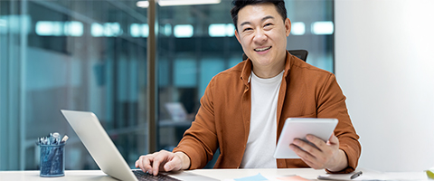 A person wearing a brown jacket and glasses sits at a desk in a modern office, smiling while holding a tablet, with an open laptop and a cup of pens nearby. Glass partitions are blurred in the background.