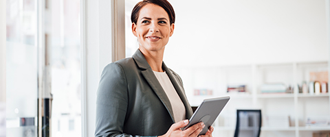 A person wearing a gray blazer stands near a large window in a bright office, smiling and holding a tablet with both hands.