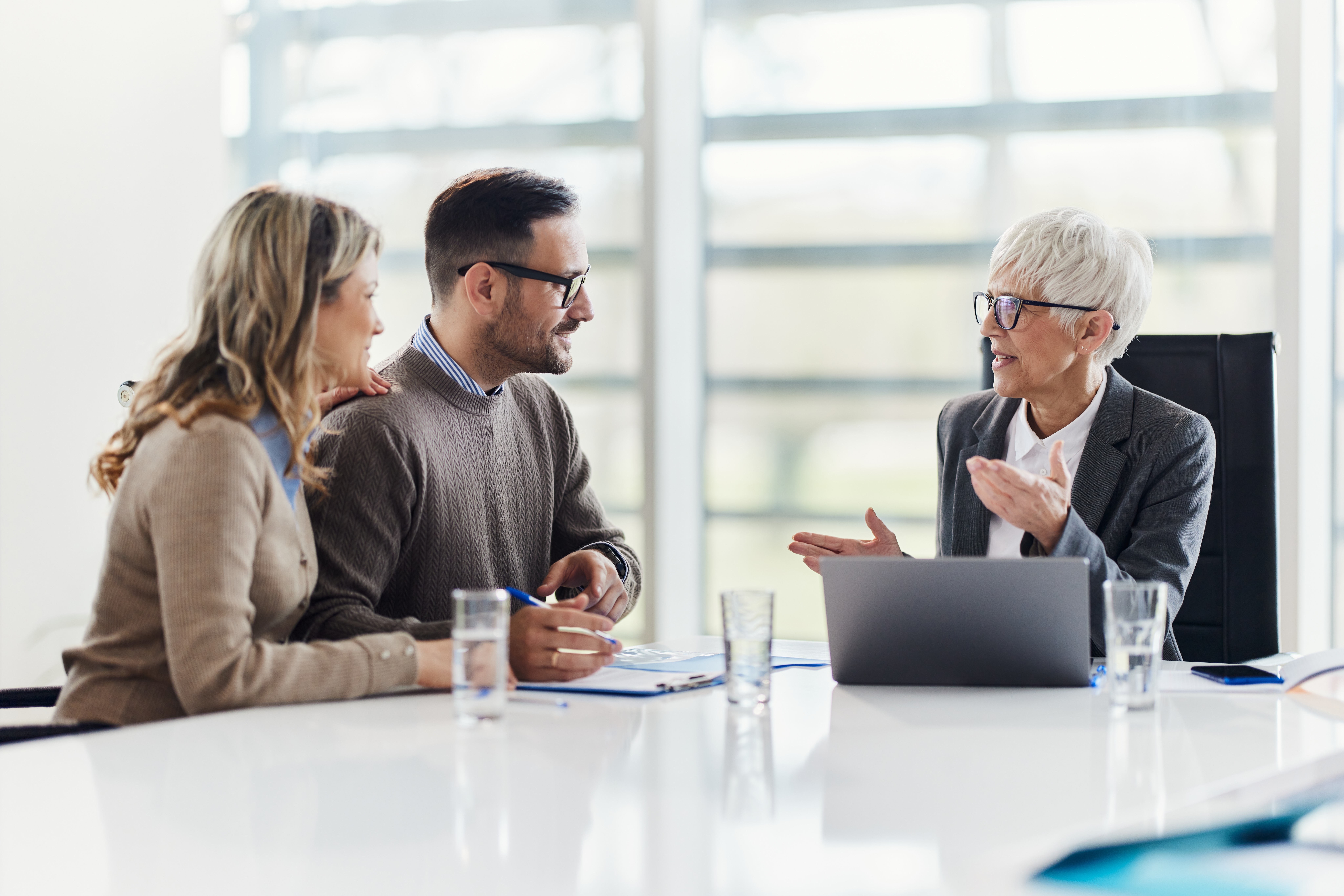 An older professional woman speaks with a younger couple across a modern office table. The three people are engaged in a serious discussion, with documents, clipboards, and glasses of water in front of them. A laptop is open in front of the advisor, suggesting a formal consultation or planning session. The bright, clean workspace and natural light create a professional and collaborative atmosphere.