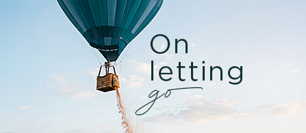 A teal hot air balloon floats in a clear sky, with its basket suspended and sand or ballast pouring out. The scene is set against a soft, pastel horizon, creating a calm and uplifting atmosphere. Overlay text reads "On letting go" in a clean, modern typeface. The overall visual suggests freedom, release, and emotional transformation.
