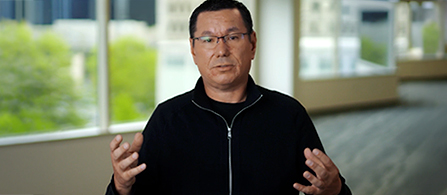 A professional in a black zip-up sweater gestures while speaking in a bright office environment. 