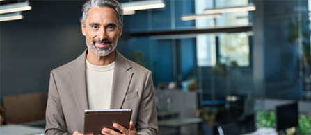 A professional holds a digital tablet in an office environment while smiling into the camera. 