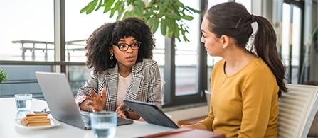 Two professionals talk while sitting at an office table with open laptops. 