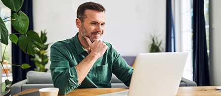 An individual sits at an office table while working on a laptop and smiling. 
