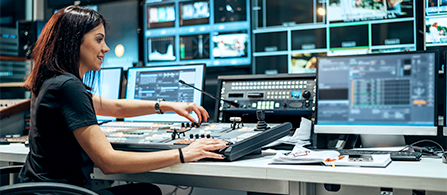Video produced in production studio adjusting dial on mixing board surrounded by video display monitors.