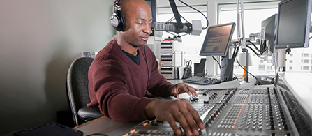  Sound editor with headphones on and speaking into microphone while adjusting dials and slides in editing booth.