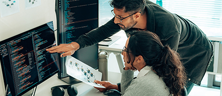  Two production engineers review flow chart while inspecting code on video monitors. 