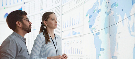 Two professionals reviewing data dashboards and a global network map on a large digital display. 