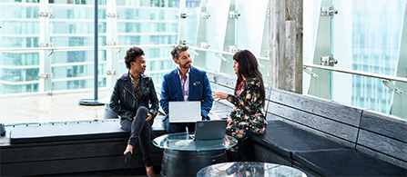 Three professionals seated in a modern office lounge, collaborating around laptops in a glass-walled workspace. 