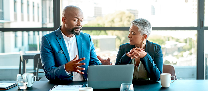Two professionals having a discussion at a conference table with a laptop in a bright office.