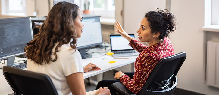 Two coworkers discussing work at a desk with computer monitors and a laptop.