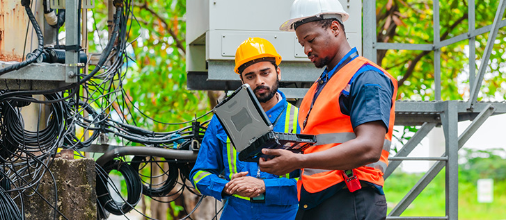 Two technicians wearing safety gear inspect network equipment at an outdoor telecommunications site.