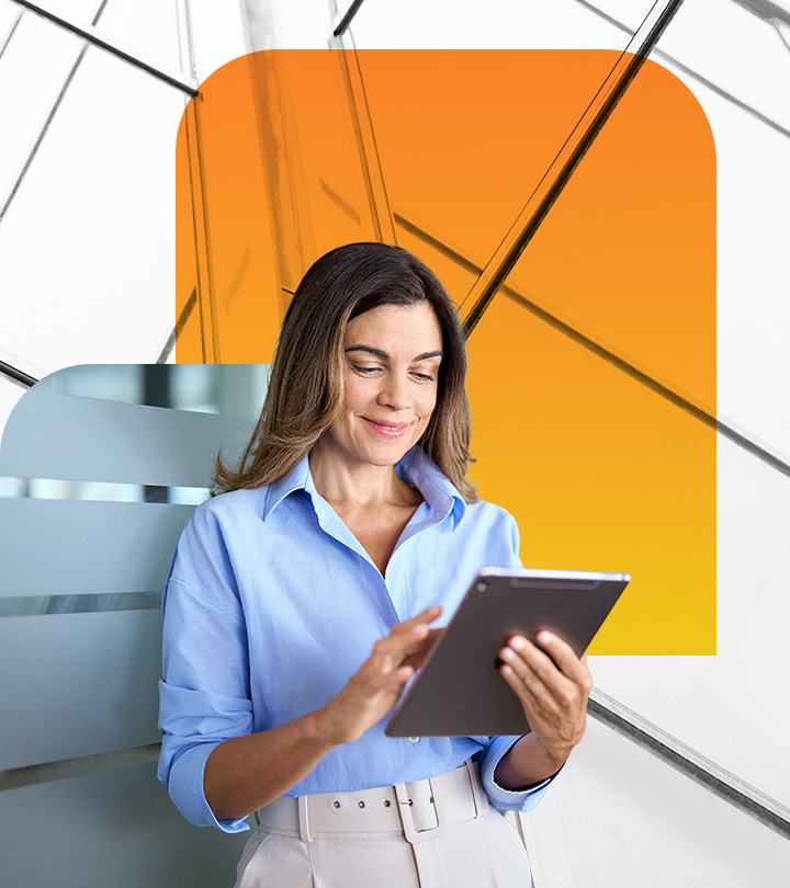 Casually dressed business person reviewing data on computer tablet standing in front of orange and gray glass office building 