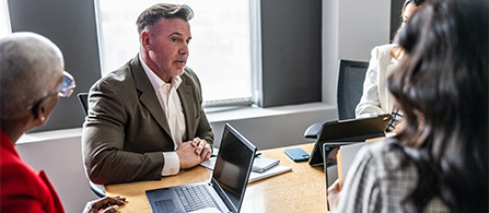 Four colleagues tightly gathered around a conference room with laptops open listening intently as one presents new ideas