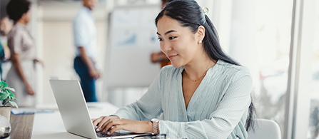 A businessperson typing on a laptop in a brightly lit open office space looks quite happy with the information on the screen.