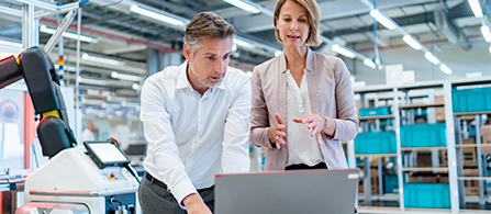 Two engineers reviewing information on computer laptop standing by a desk in a robotic manufacturing room. 