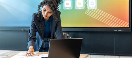 A person with curly hair in a dark blazer over a teal top leans forward over a table, looking down at papers while touching them with one hand. A laptop sits open in front of them and a large display showing a colorful gradient with icons glows behind them.