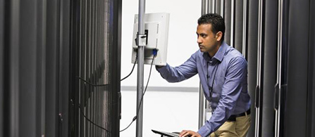 A person in a blue shirt and lanyard badge stands in a dark data center aisle, leaning forward to work on a monitor mounted on an open server rack. Tall black server cabinets with indicator lights line both sides.