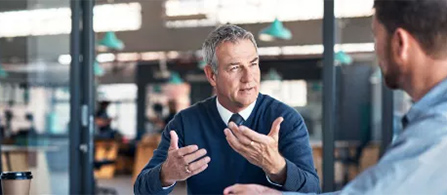 A person with gray hair wearing a navy sweater and tie gestures with both hands while speaking to another person seen from behind across a table, in an open office with hanging pendant lights.