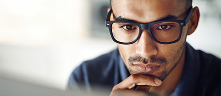 A close-up of a person wearing dark-framed glasses resting their chin on their hand with a thoughtful, focused expression, against a softly blurred dark background.