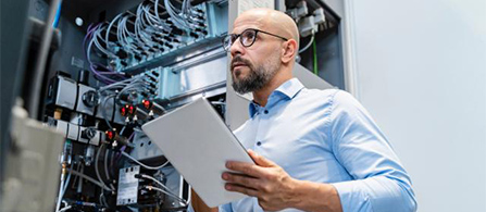 A person with a beard and glasses, wearing a light blue shirt, holds a tablet while looking up at a wall of network cabling and equipment in a data center or wiring closet.