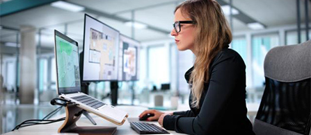 A person with glasses and long hair, wearing a dark top, sits at a desk working on a laptop raised on a stand beside an external monitor displaying maps and data. The setting is a bright modern office with windows behind.