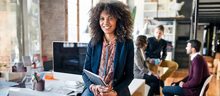 A person in a dark blazer sits on the edge of a desk holding a tablet and smiling, in a bright open office. Several colleagues are gathered in a lounge area behind them.