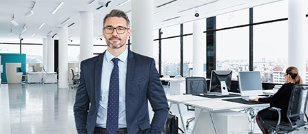 A person with gray hair and glasses wearing a navy suit and tie stands smiling in a bright, open office with large windows. A colleague works at a desk in the blurred background.