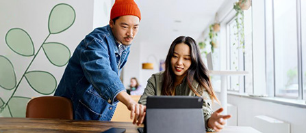 Two people stand side by side at a desk in a bright café-style workspace, looking at a tablet held by one of them. One wears a red knit cap and a denim jacket; the other smiles toward the screen. Plants and large windows fill the background.