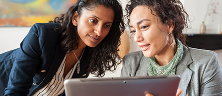 Two people sit close together looking at a tablet held between them. One in a dark blazer leans in and looks at the screen; the other in a gray blazer with a green scarf smiles toward the device. A blurred office setting is behind them.