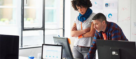 Two people stand at a desk in a bright office looking at a monitor. One with curly hair wearing a blue scarf and lanyard stands with arms crossed; the other in a plaid shirt points toward the screen. Whiteboards and charts fill the background.