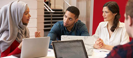 Three people sit together at a table in a bright collaborative space, engaged in conversation. One wearing a headscarf gestures while speaking; the other two listen attentively. Laptops rest open on the table and red accents fill the background.