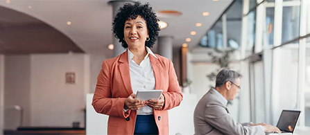 A person with curly hair in a coral blazer over a white top stands in a bright lobby area, smiling toward the camera and holding a tablet. A colleague works at a desk in the softly blurred background.