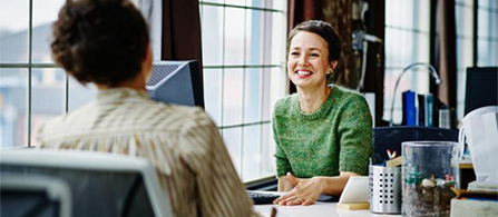 Two people sit across from each other at desks in a bright office near large windows. One in a green sweater smiles warmly toward the other, who is seen from behind. Pens and a small container sit on the desk.