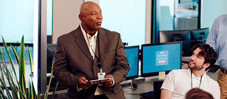 A person in a tan sport coat holding a tablet stands and speaks in a busy open office, facing a seated colleague who looks up and listens. Monitors, a plant, and other colleagues fill the bright background.