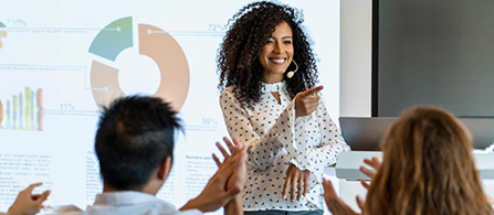 A person with curly hair in a white patterned blouse stands at the front of a conference room, smiling and gesturing while presenting to a seated audience. A large projection screen showing charts is visible behind them.