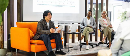 Three people sit in a casual panel setting on orange armchairs and chairs in a bright space. One in a patterned blazer holds papers and speaks; two others sit listening. A projection screen with text is partially visible behind them.