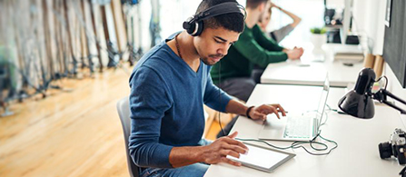 A person in a blue sweater and headphones sits at a desk using a tablet and laptop in a bright open office. A colleague works at another desk in the background.
