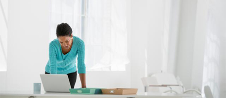 A person in a teal sweater stands leaning over a desk working on a laptop in a bright, minimal white room with a window and an open folder beside them.