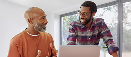 Two people talk and smile at each other across a laptop in a bright office. One wears an orange sweater and gestures while speaking; the other wears a plaid shirt and glasses, leaning on a desk.