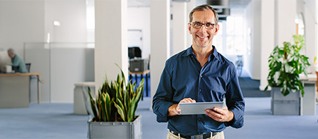 A person with gray hair and glasses, wearing a blue shirt, stands smiling and holding a tablet in a bright open office with potted plants and a colleague working in the background.