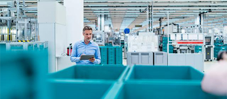A person in a light blue shirt stands holding a tablet on a factory or warehouse floor, surrounded by large teal bins and manufacturing equipment receding into the background.