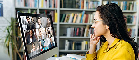 A person with long dark hair in a yellow top sits at a desk in profile, resting a hand on their chin and looking at a large monitor showing a video call with multiple participants in a grid. Bookshelves fill the background.