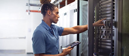 A person with hair in an updo wearing a blue polo shirt stands in a data center aisle, reaching into an open server rack while holding a tablet in the other hand. Rows of white equipment cabinets line the background.