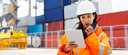 A person wearing a white hard hat and an orange high-visibility jacket stands outdoors at an industrial port, holding a tablet in one hand and a radio in the other while looking down at the screen. Shipping containers and equipment fill the background.
