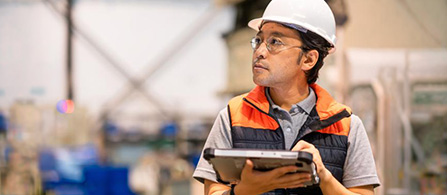 A person wearing a white hard hat, orange safety vest, and glasses stands in an industrial facility, holding a tablet and looking upward with a focused expression. Equipment and machinery fill the blurred background.