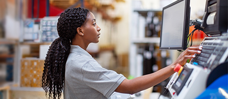A person with braided hair in a gray shirt stands at an industrial control panel in a warehouse setting, looking at a monitor and operating controls with a focused expression.