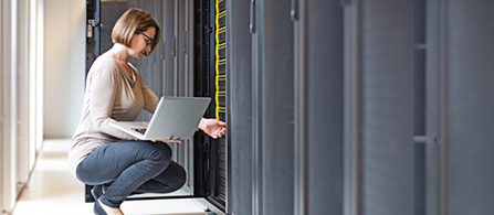 A person wearing glasses crouches beside an open server rack in a data center corridor, holding a laptop and examining the equipment with a focused expression.