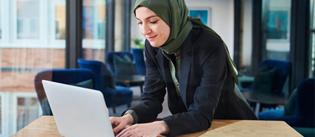 A person wearing a green head covering and a black blazer sits at a wooden table in a modern office, smiling while typing on a laptop. Blue chairs and large windows are visible in the background.
