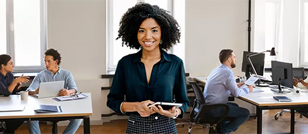 A person in a dark teal shirt stands in the foreground of a busy open-plan office, smiling and holding a tablet. Two other people are working at desks in the background.